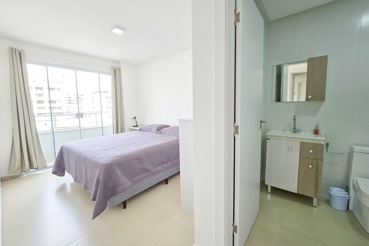 Een slaapkamer met een bed, nachtkastje, raam met gordijnen en een badkamer met wastafel en spiegel.
