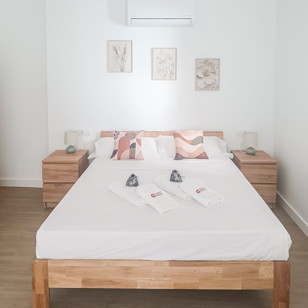 Una cama bien hecha con una colcha blanca, dos mesitas de noche de madera con lámparas y tres cuadros enmarcados en la pared.