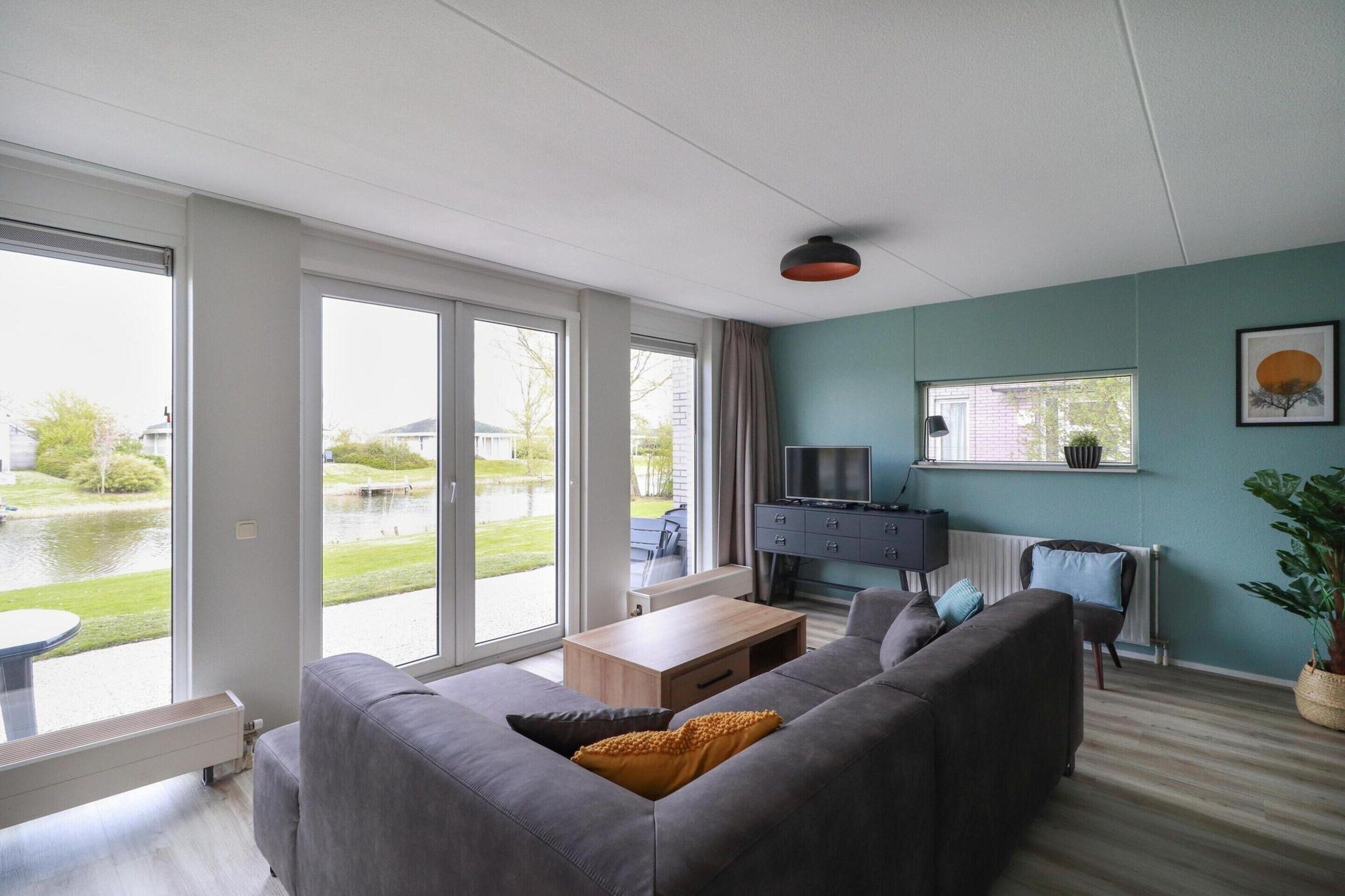 Un salon moderne avec un canapé gris, une table basse en bois et un téléviseur posé sur un meuble noir. Une grande fenêtre offre une vue sur un plan d’eau et la verdure environnante.