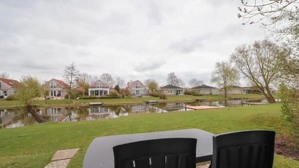 Blick auf ein Seeufergrundstück mit Tisch und Stühlen, einen Teich und Häuser im Hintergrund.