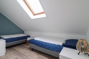 Ein kleiner Raum mit schrägem Dach, zwei Betten, einem kleinen Hocker und einem Korbstuhl.
