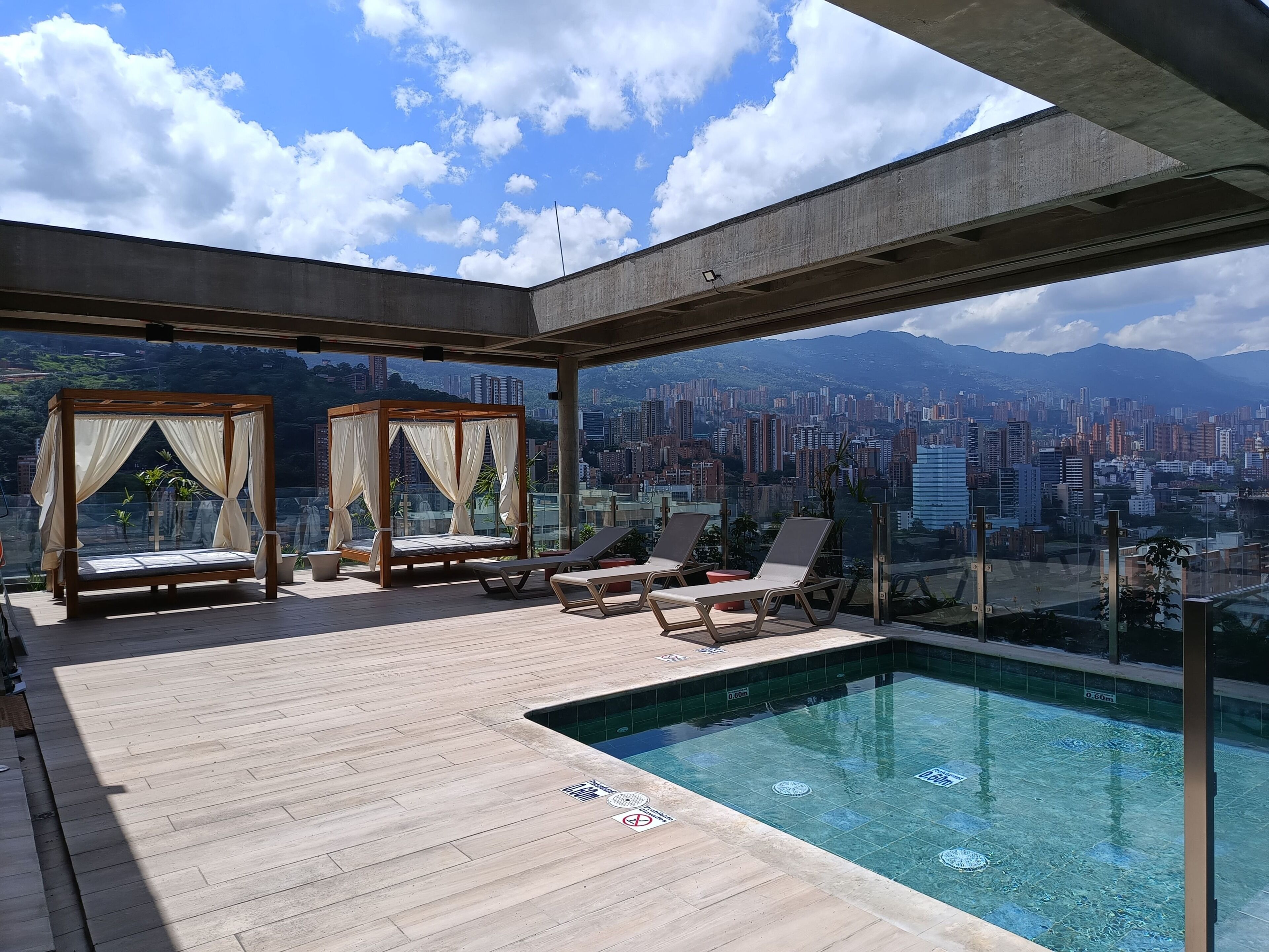 A rooftop pool area with a bed and lounge chairs, a view of a cityscape, and a clear sky.