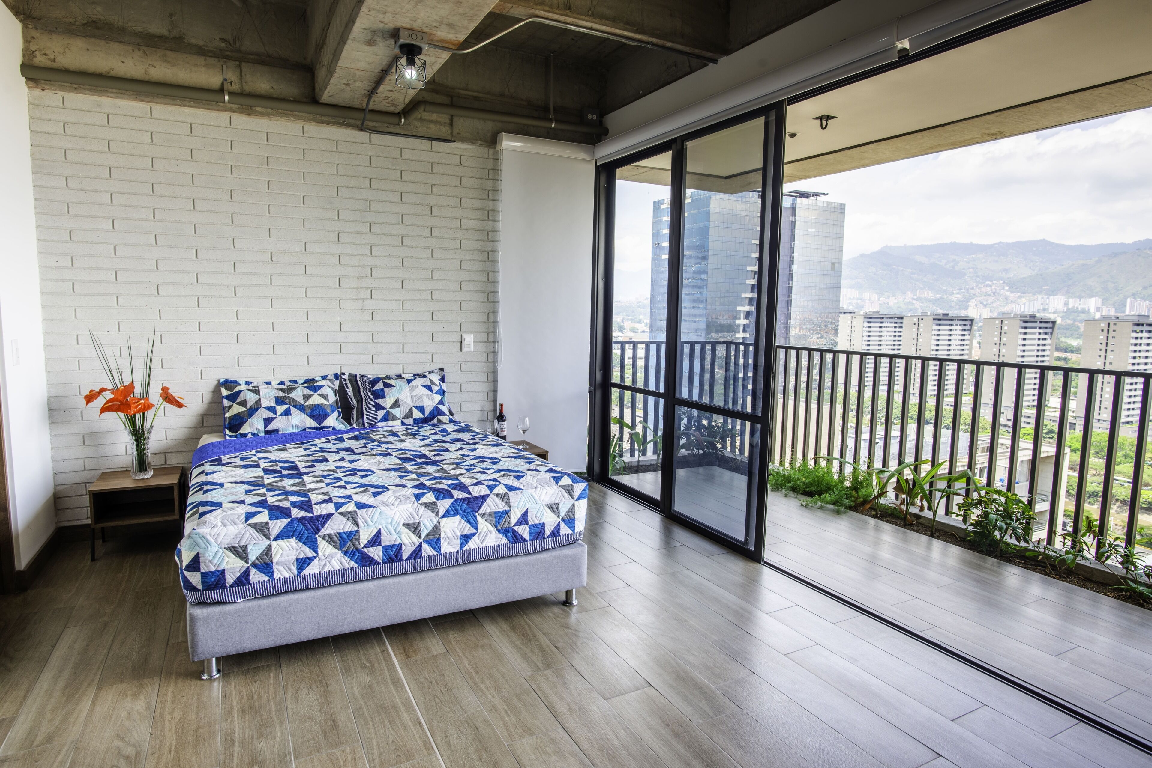 Une chambre avec un grand lit, un mur en briques blanches, un plancher en bois et un balcon offrant une vue sur la ville.