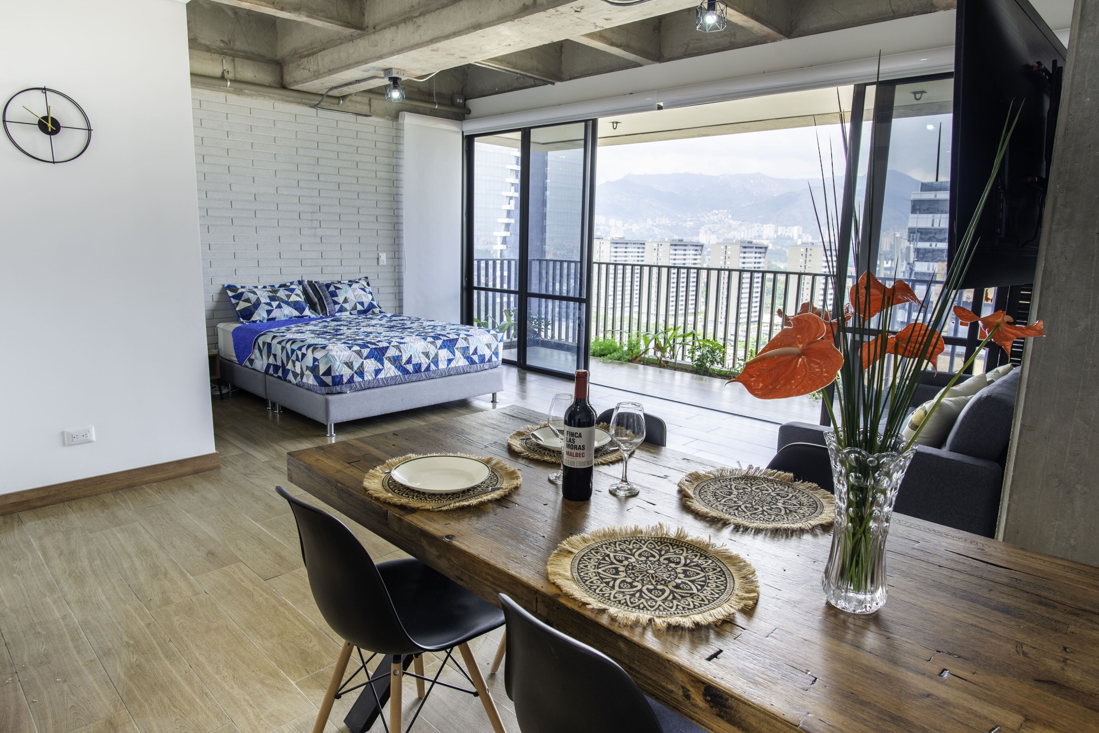 Eine moderne Wohnung mit Essbereich, einem Bett mit gemusterten Betttextilien und einem Balkon mit Blick auf die Stadt.