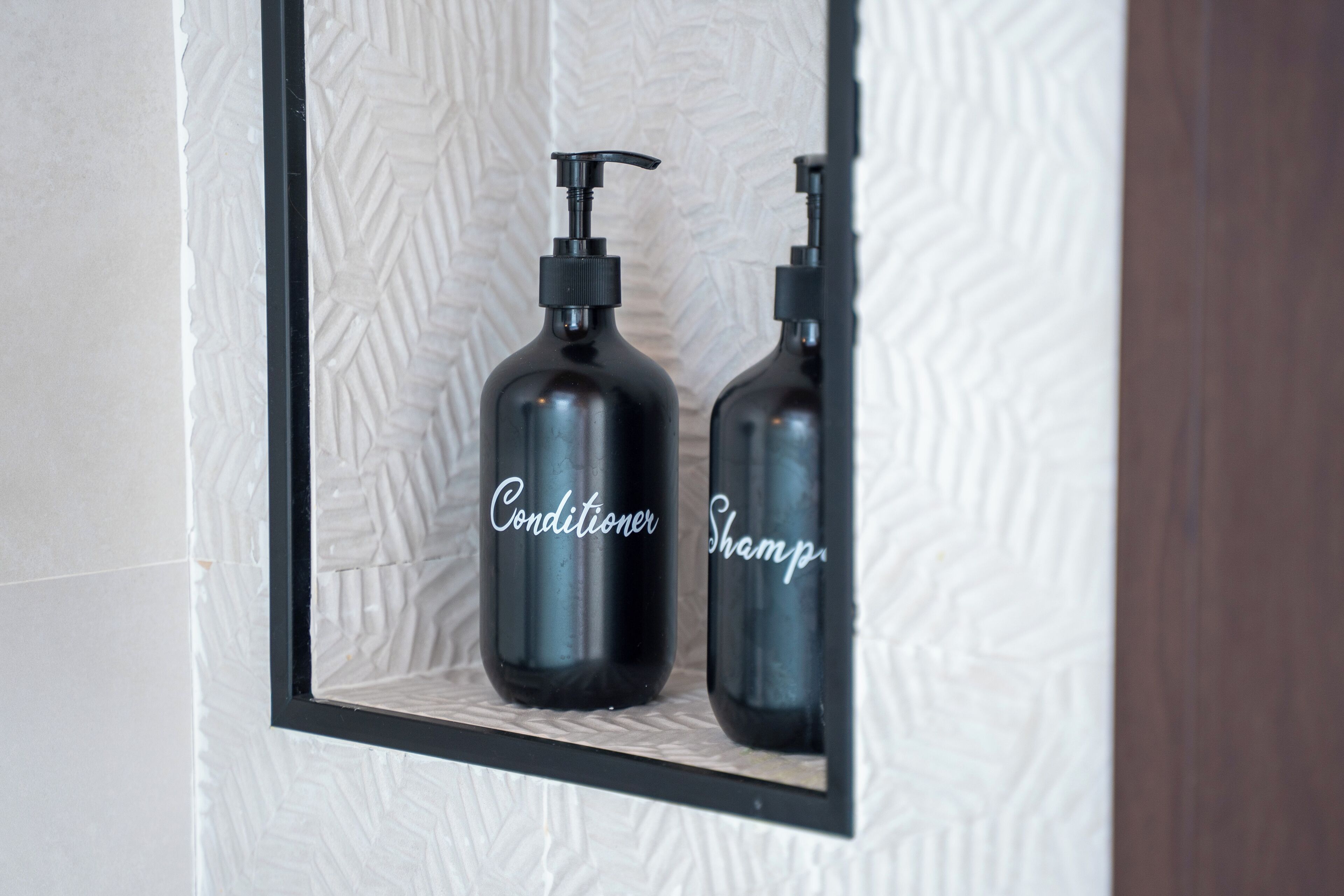 Un miroir reflète deux bouteilles noires portant les étiquettes « Après-shampoing » et « Shampoing », le tout se trouvant sur un mur à texture.