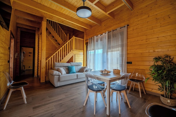 A wooden cabin interior with a staircase, a sofa, a dining table with chairs, and a potted plant.