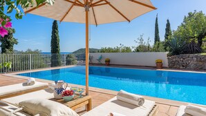 Área da piscina com espreguiçadeiras, uma mesa com jarra e frutas, e vista para o mar.