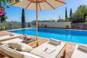 Ein Poolbereich mit Liegestühlen, einem Tisch mit einem Krug und Früchten sowie Blick aufs Meer.