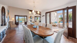 Uma sala de jantar ampla com uma grande mesa de madeira e cadeiras, um lustre e vista para o exterior através de grandes portas de vidro deslizantes.
