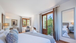 Quarto de hotel com uma cama grande, área de estar com uma poltrona e mesa, varanda com vista e um espelho grande.