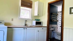 Une cuisine de taille réduite, dotée d’un évier blanc, d’un four à micro-ondes et d’une fenêtre avec des stores.
