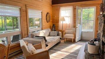 A cozy cabin interior with a sofa, chairs, a fireplace, and large windows.