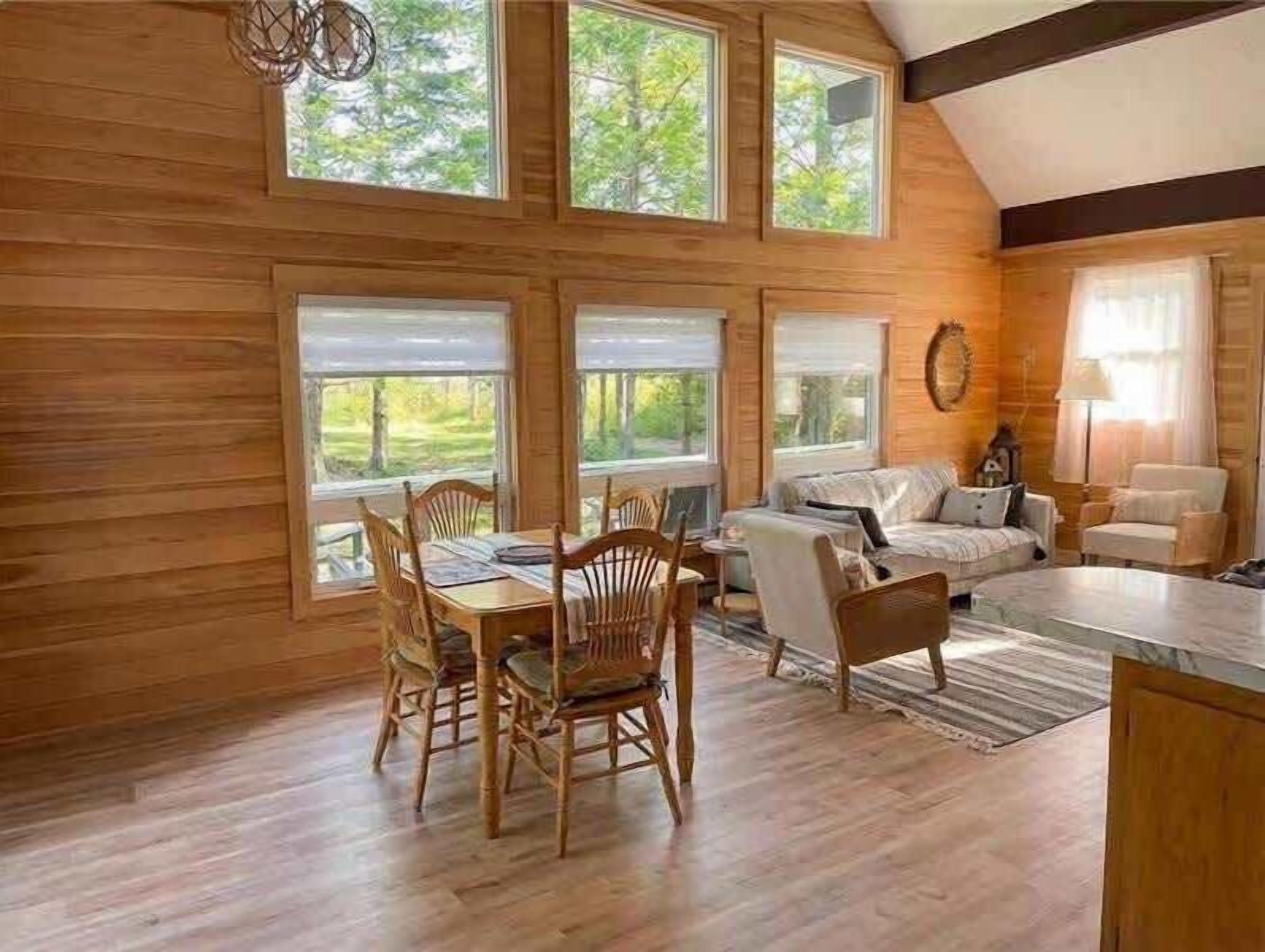A wooden interior with a dining table and chairs, a sofa, and a window with a view of greenery.
