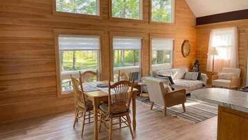 A wooden interior with a dining table and chairs, a sofa, and a window with a view of greenery.