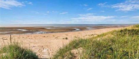 Ein sandiger Strand mit einem hölzernen Promenadenweg, der zum Wasser führt, unter blauem Himmel.