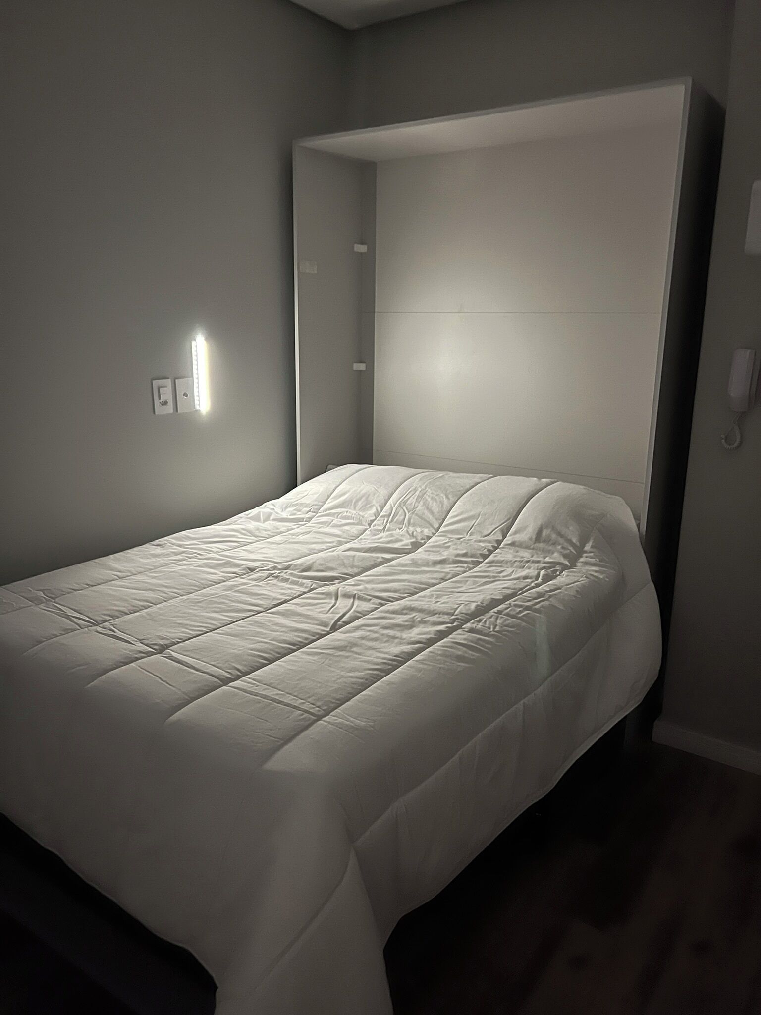 A single bed with a headboard in a room with a light switch and a wall socket.