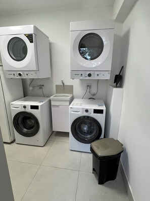 A laundry room with two front-loading washing machines, a sink, and a trash bin. - Loft4 Brooklin Berrini Vila Olímpia Airport CGH (São Paulo)