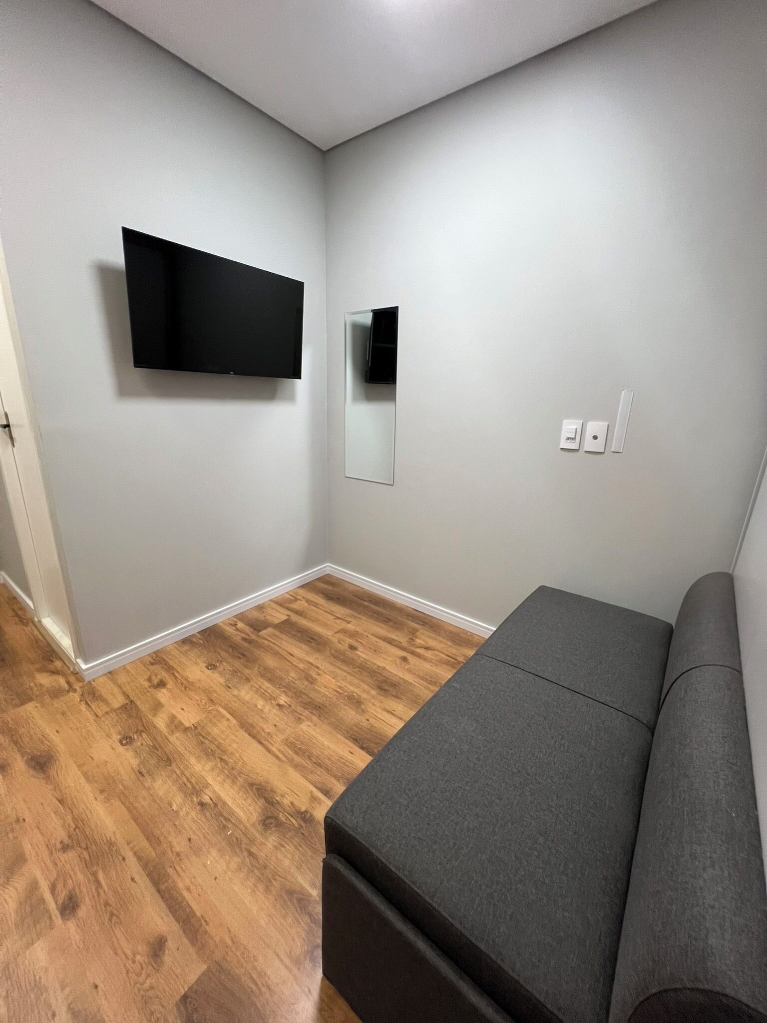 A room with a grey sofa, a flat-screen TV mounted on the wall, and a wooden floor.