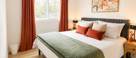 Un dormitorio con cama, cortinas, mesita de noche, lámpara y ventana.