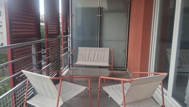 Een balkon met een metalen hekwerk, een glazen deur en twee witte stoelen.