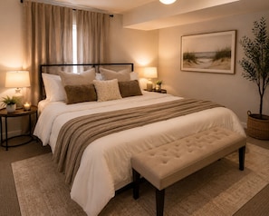 Quarto com cama, mesas de cabeceira, apoio para luminária, um banquinho, luminária, planta e um quadro na parede.