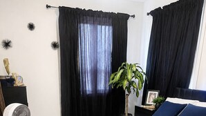 A bedroom with a bed, a bedside table, a fan, a plant, and a window with curtains.