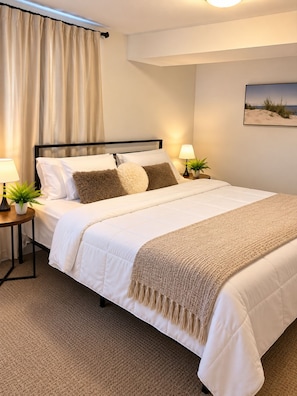 Ein ordentlich bezogenes Bett mit einer weißen Daunendecke und mehreren Kissen, ein Nachttisch mit einer Lampe und einer Pflanze sowie ein Strandbild an der Wand.