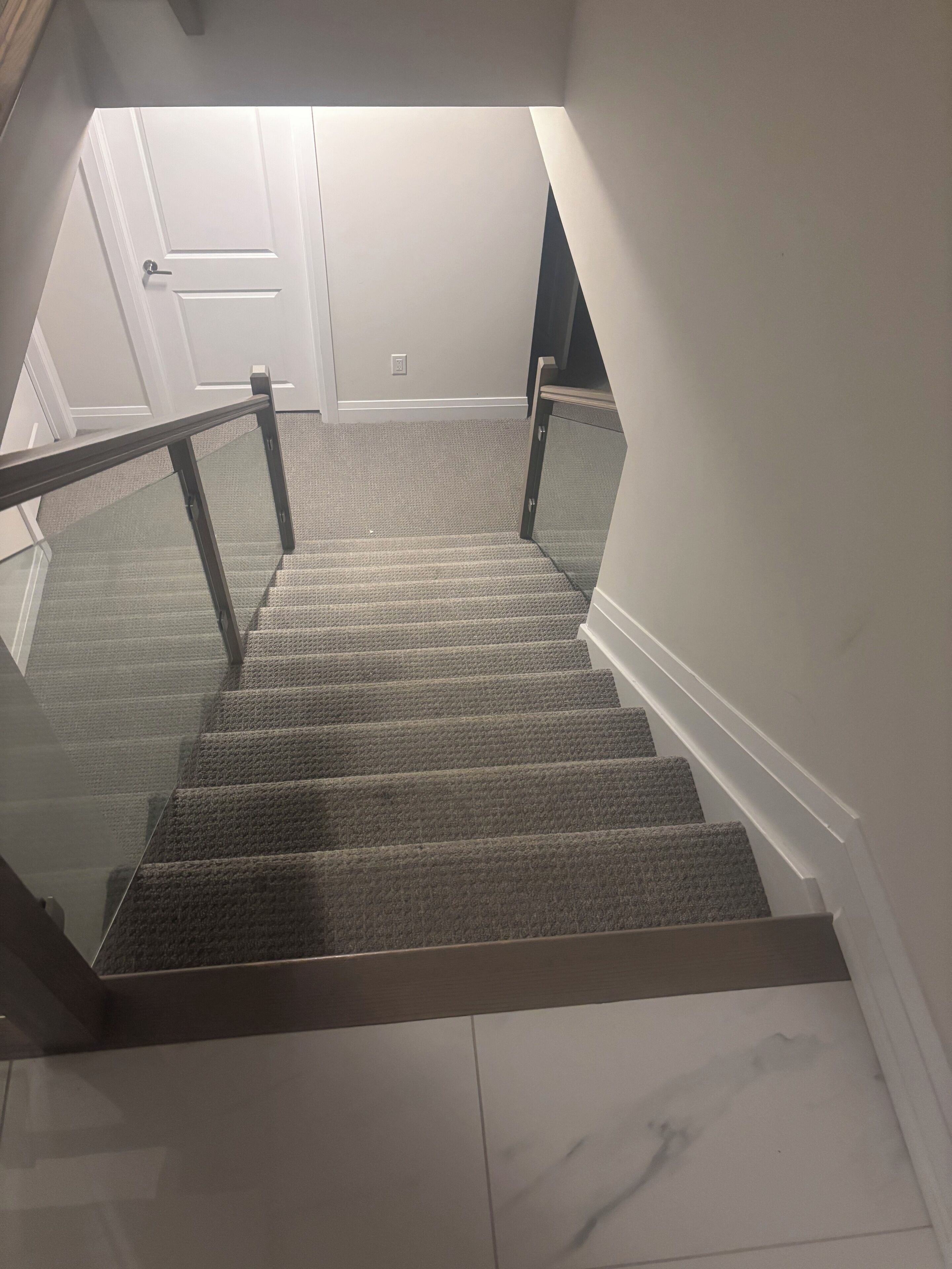 Una escalera alfombrada con barandilla de cristal, paredes blancas y una puerta en la parte superior.