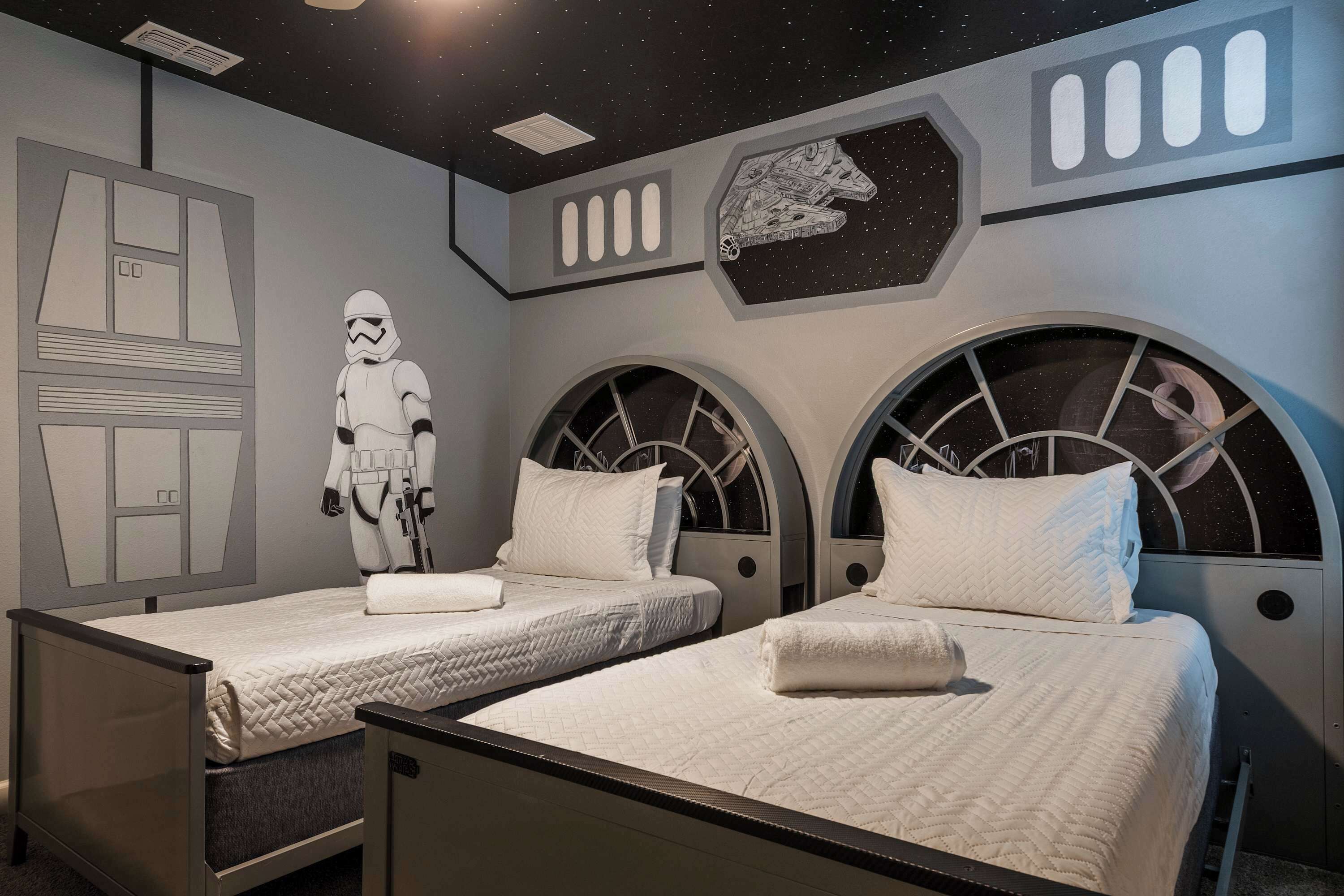 A room with two beds, each with a starry night sky design on the headboard, a wall with a space-themed mural, and a Stormtrooper figure.