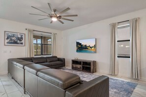 Ein modernes Wohnzimmer mit einem Ecksofa, Deckenventilator, Flachbildfernseher und einem großen Fenster mit Vorhängen.