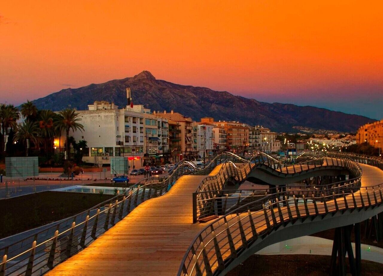 A curved pedestrian bridge with railings, leading to a waterfront area with buildings and palm trees.