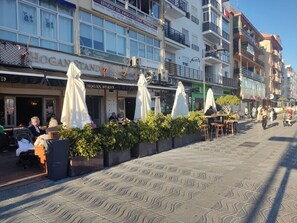 A street with outdoor seating, a restaurant named 'Hogan Stand,' and multi-story buildings with balconies. - RicosRincones · Ocean View Apartment (San Pedro Alcántara)