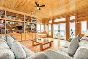 Ett rymligt vardagsrum med ett träpanelat tak, en stor bokhylla, en tv och utsikt över stranden genom glasdörrarna.
