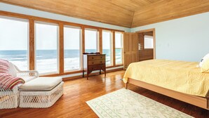 Quarto com uma grande janela com vista para o oceano, piso de madeira, uma cama com colcha, uma cadeira e uma cômoda.