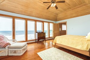 Ein Schlafzimmer mit großem Fenster und Meerblick, Holzboden, einem Bett mit Steppdecke, einem Sessel und einer Kommode.