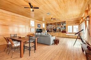 A spacious living area with a wooden ceiling, a sofa, a dining table with chairs, and a bookshelf filled with books. - Steps from the Beach, Pet Friendly, Warm Interior with Great Views (Galveston)
