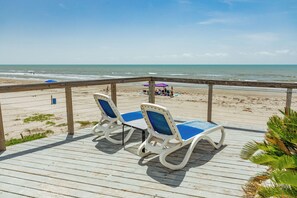 A wooden deck with two lounge chairs and a small table, overlooking a beach with a clear view of the ocean. - Steps from the Beach, Pet Friendly, Warm Interior with Great Views (Galveston)
