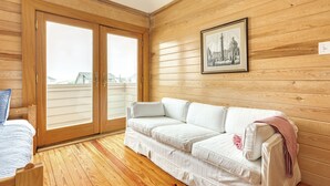 A room with a wooden floor, a white sofa, a bed with a blue quilt, and a large window with a view of houses.
