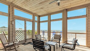 A covered outdoor seating area with wooden flooring, metal-framed chairs, and a rocking chair. Large windows provide views of the beach and ocean.