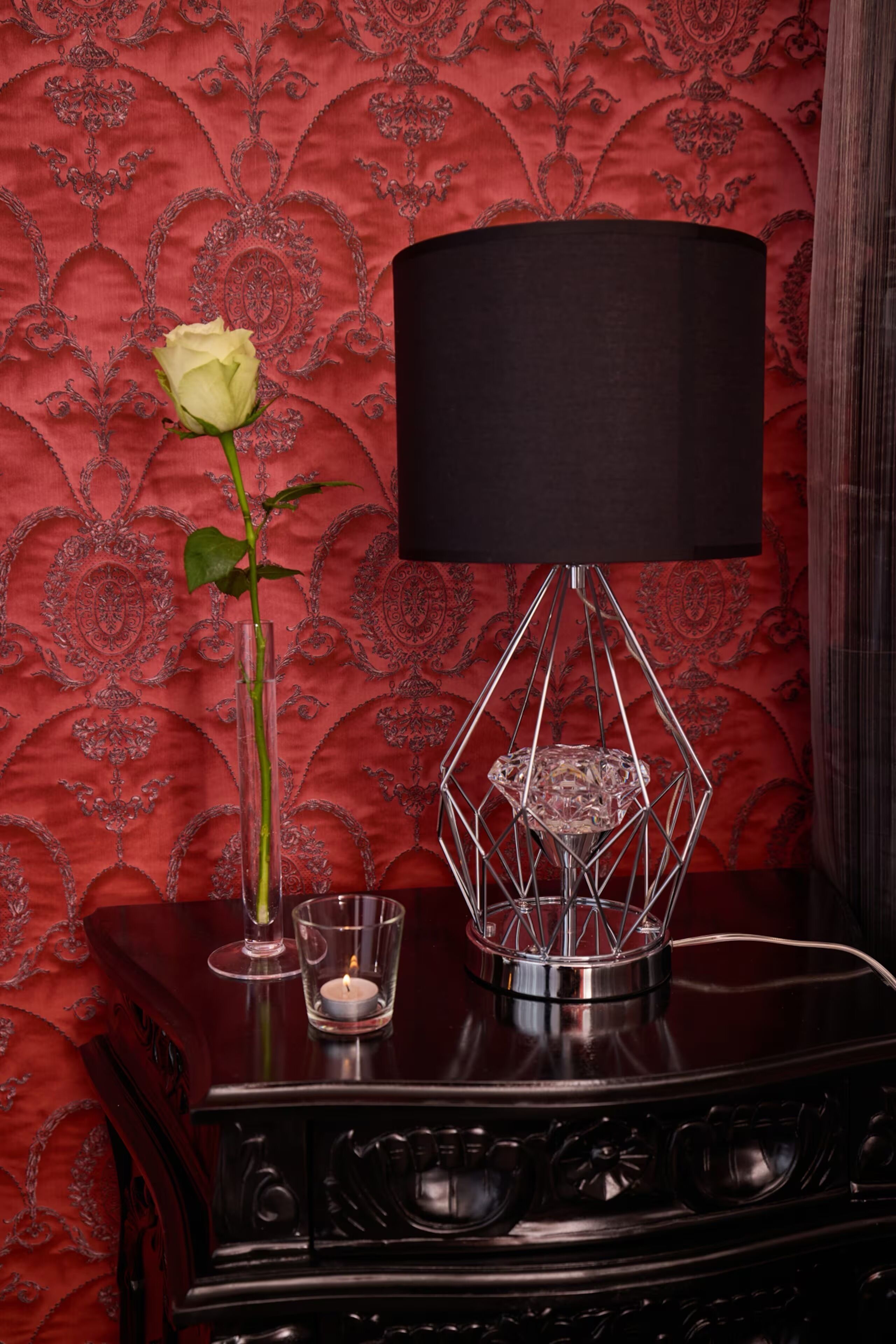 A table with a black lamp, a crystal vase, a lit candle, and a white rose.