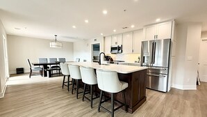 A modern kitchen with a central island, bar stools, stainless steel appliances, and a dining area with a table and chairs. - New Spacious 5-bedroom condo in the heart of fabulous Dewey Beach (Dewey Beach)