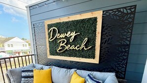 A balcony with a grey sofa, yellow cushions, and a decorative rug. A sign reads 'Dewey Beach'. - New Spacious 5-bedroom condo in the heart of fabulous Dewey Beach (Dewey Beach)