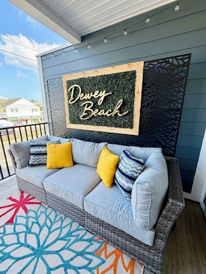 Ein Balkon mit einer grauen Couch, gelben Kissen und einem dekorativen Teppich. Ein Schild zeigt 'Dewey Beach'.