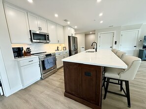 A modern kitchen with white cabinetry, a central island, and a dining area with a bar stool. - New Spacious 5-bedroom condo in the heart of fabulous Dewey Beach (Dewey Beach)