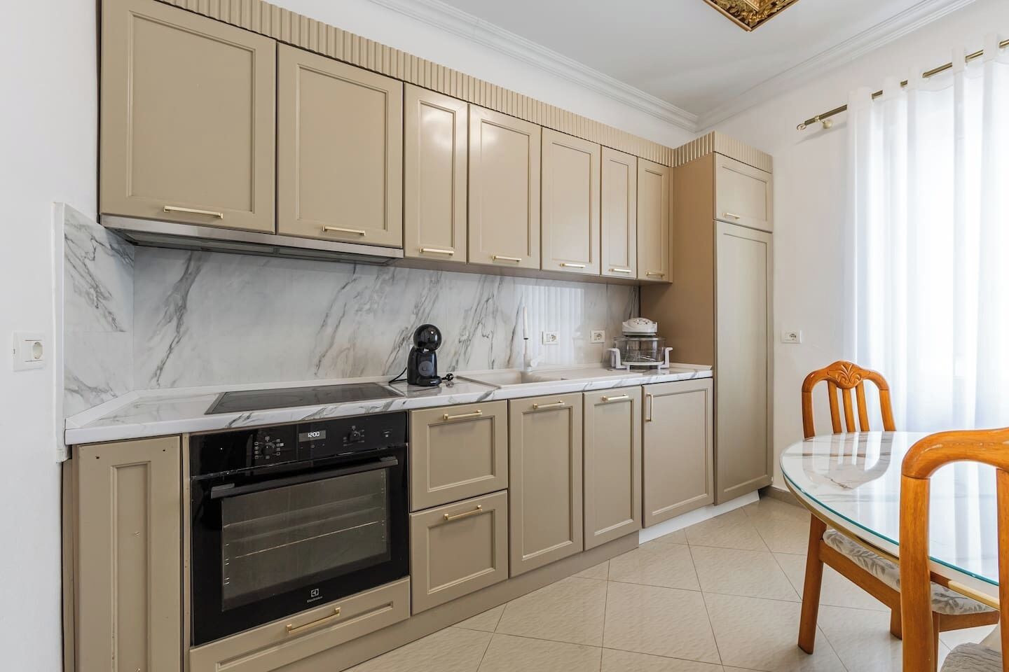 A modern kitchen with beige cabinets, a built-in oven, and a glass dining table with chairs.