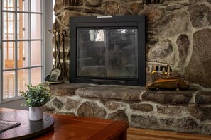 A stone fireplace with a glass door, a potted plant on the mantel, and a wooden table in the foreground. - Mountain Cabin with Arcade, Hot Tub & Fire Pit (Berkeley Springs)