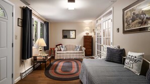 A cozy room with a bed, a sofa, a small table, and a chair. There is a window with curtains, a lamp, and a painting on the wall. - Right Mtn Attitude · Arcade, Hot Tub & Fire Pit (Berkeley Springs)