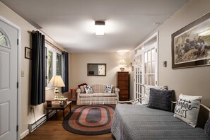 A cozy room with a bed, a sofa, a small table, and a chair. There is a window with curtains, a lamp, and a painting on the wall. - Right Mtn Attitude · Arcade, Hot Tub & Fire Pit (Berkeley Springs)