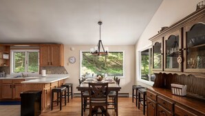 A spacious kitchen with wooden cabinets, a dining area with a large window, and a view of greenery outside. - Right Mtn Attitude · Arcade, Hot Tub & Fire Pit (Berkeley Springs)