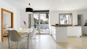 A modern kitchen with a central island, a dining area with a table and chairs, and a large sliding glass door leading to a patio. - Beachfront Villa With Pool, Puerto Pollenca (Pollença)